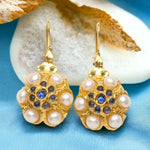 Gold flower shaped earrings with pearl like beads and blue stones on a blue background with white stone