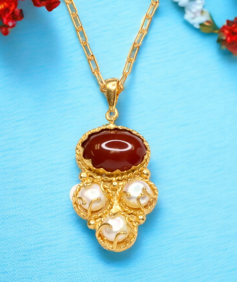 Gold colored necklace with a red stone and pearl-like accents on a gold elongated link chain on a blue background