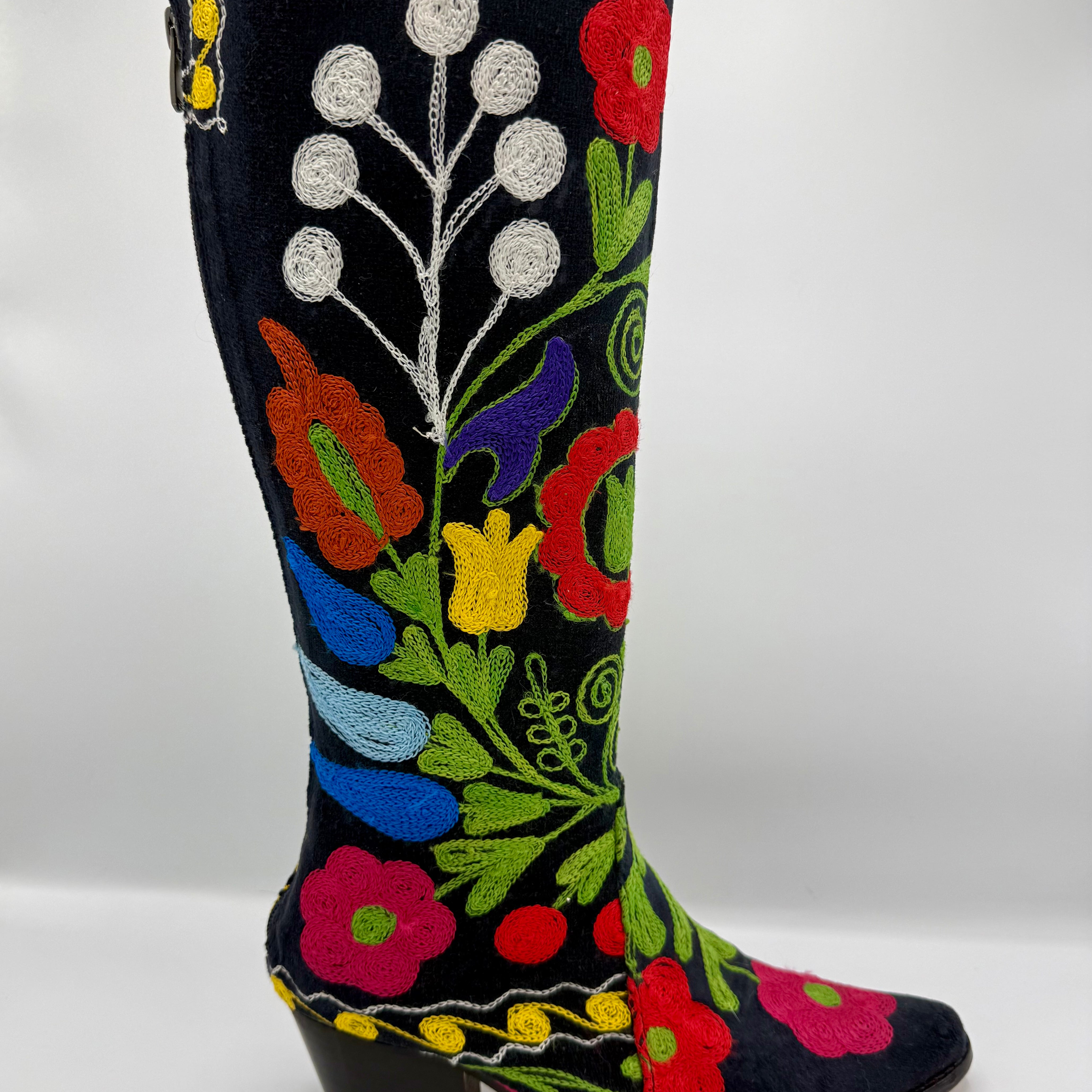 A black suede cowboy boot with vibrant multicolor floral embroidery, featuring a low black block heel