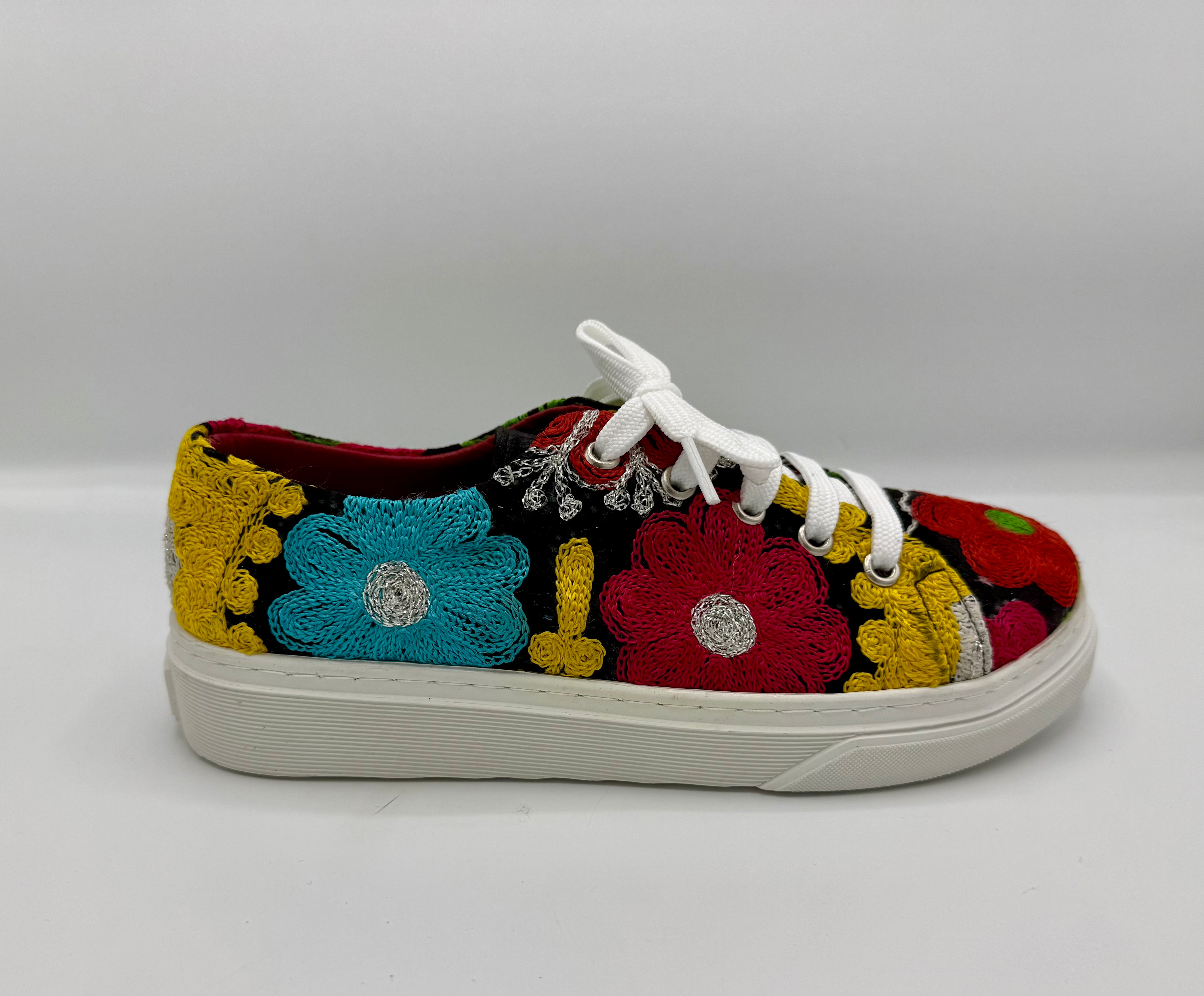 A low-top canvas sneaker featuring vibrant, multicolor floral embroidery in red, blue, yellow, and green on a black background, with white laces and a white rubber sole.