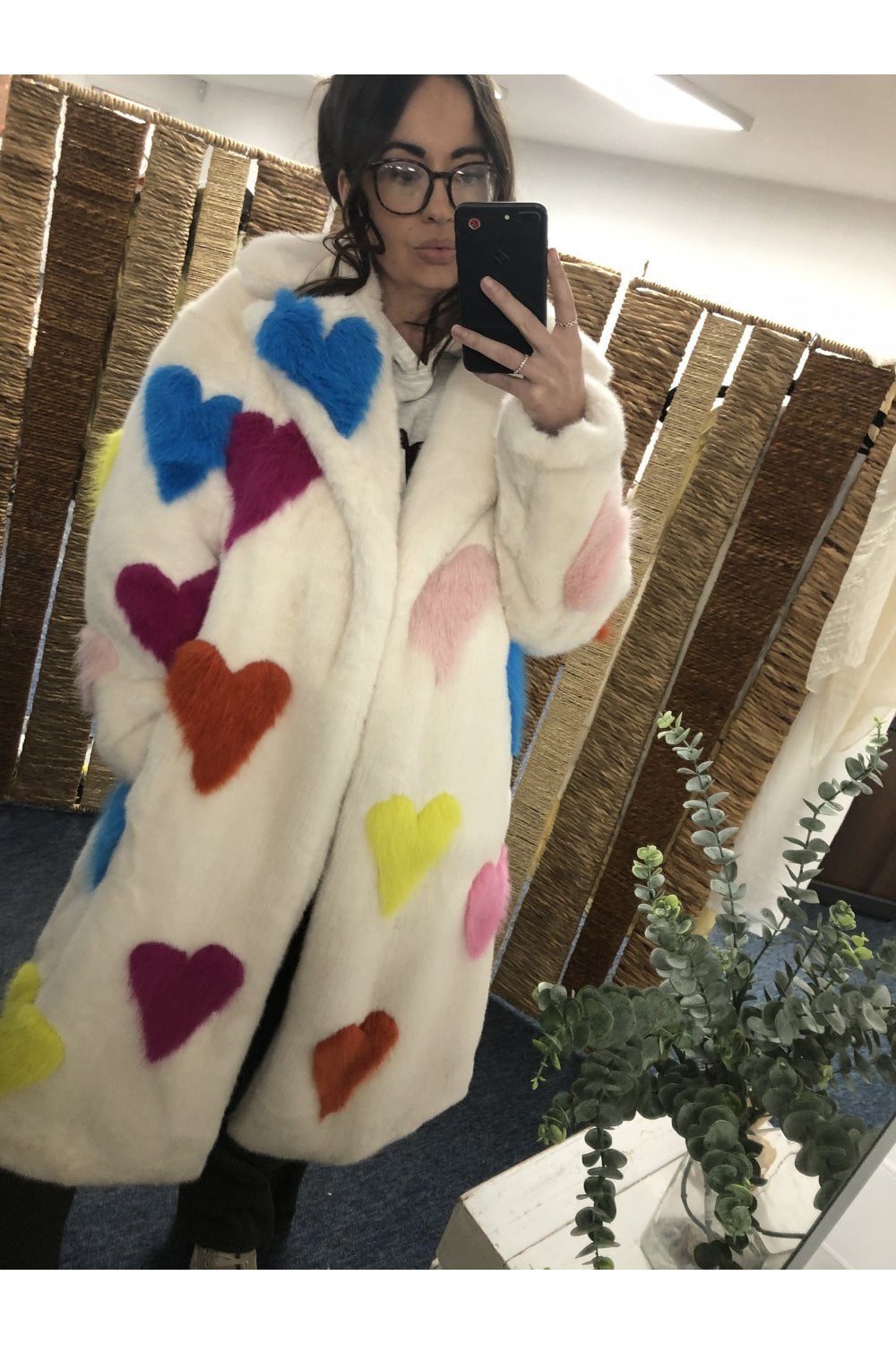 Person wearing a white coat with colorful heart patterns taking a mirror selfie.
