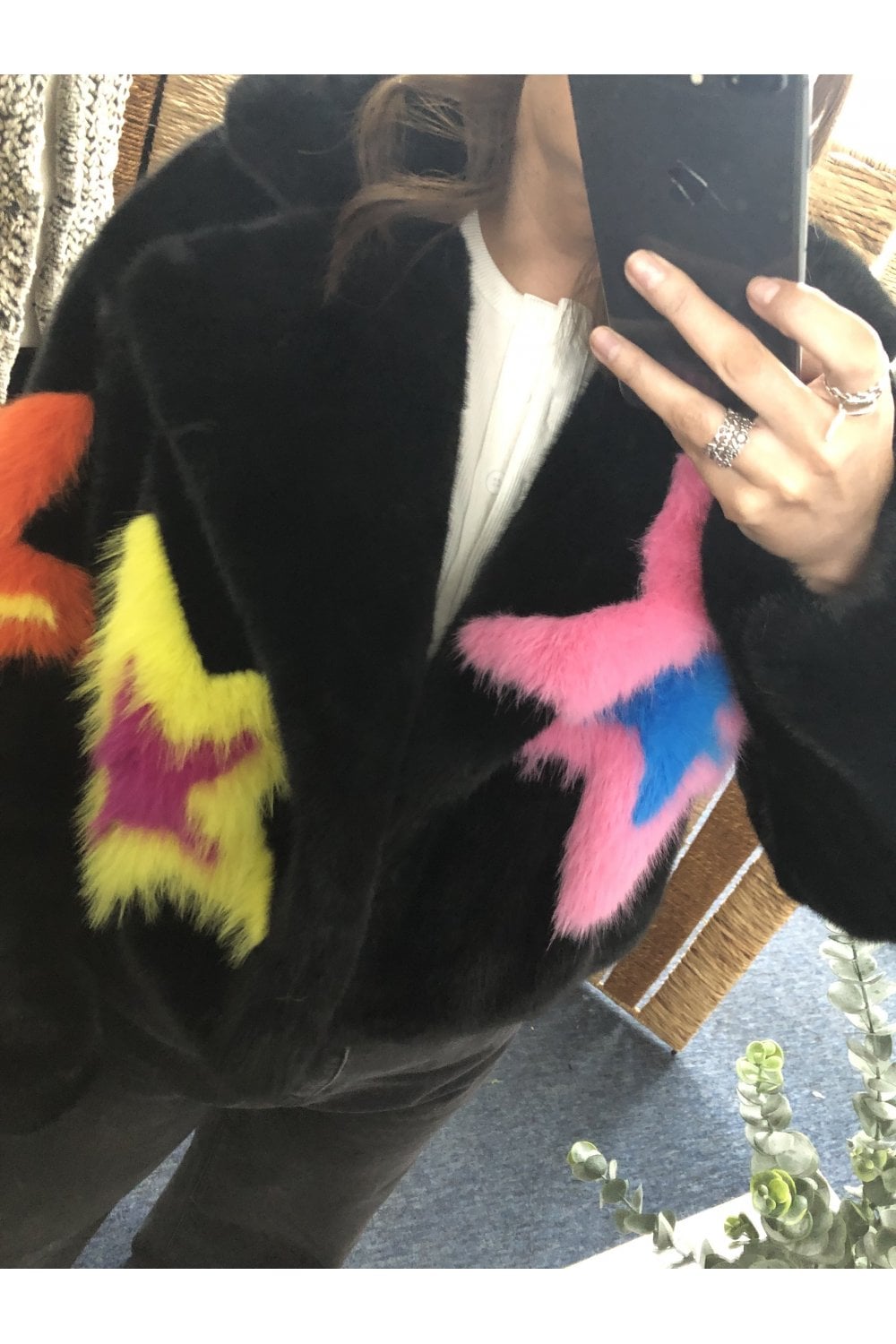 Person wearing a black coat with colorful fur star shaped patches, taking a mirror selfie.