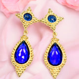 Gold colored earrings with blue teardrop stones on a pink fabric background