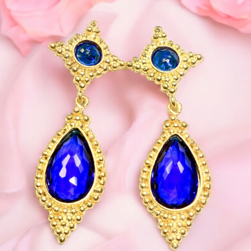 Gold colored earrings with blue teardrop stones on a pink fabric background