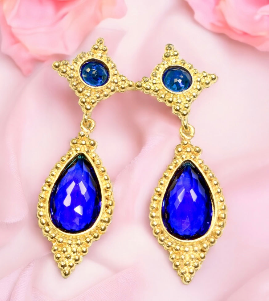 Gold colored earrings with blue teardrop stones on a pink fabric background