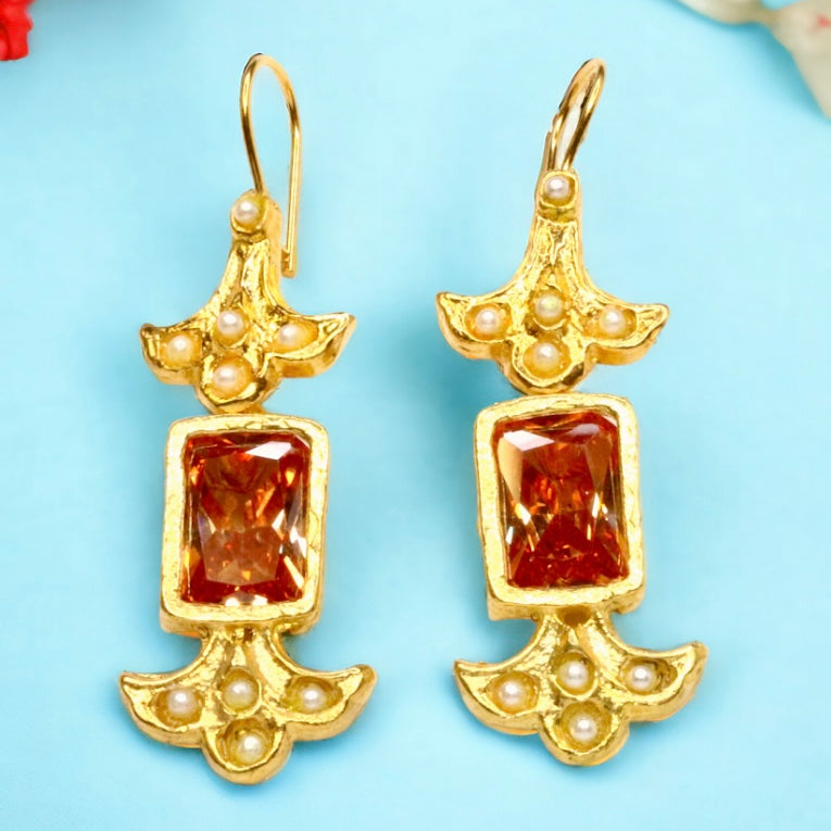 Gold colored earrings with orange stones and pearl like beads on a light blue background