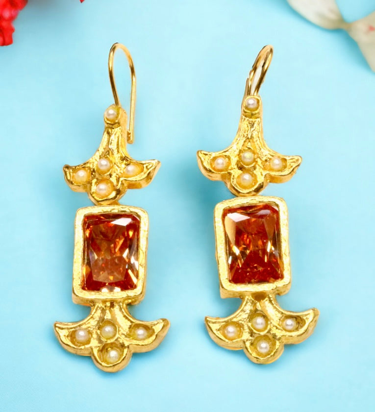 Gold colored earrings with orange stones and pearl like beads on a light blue background