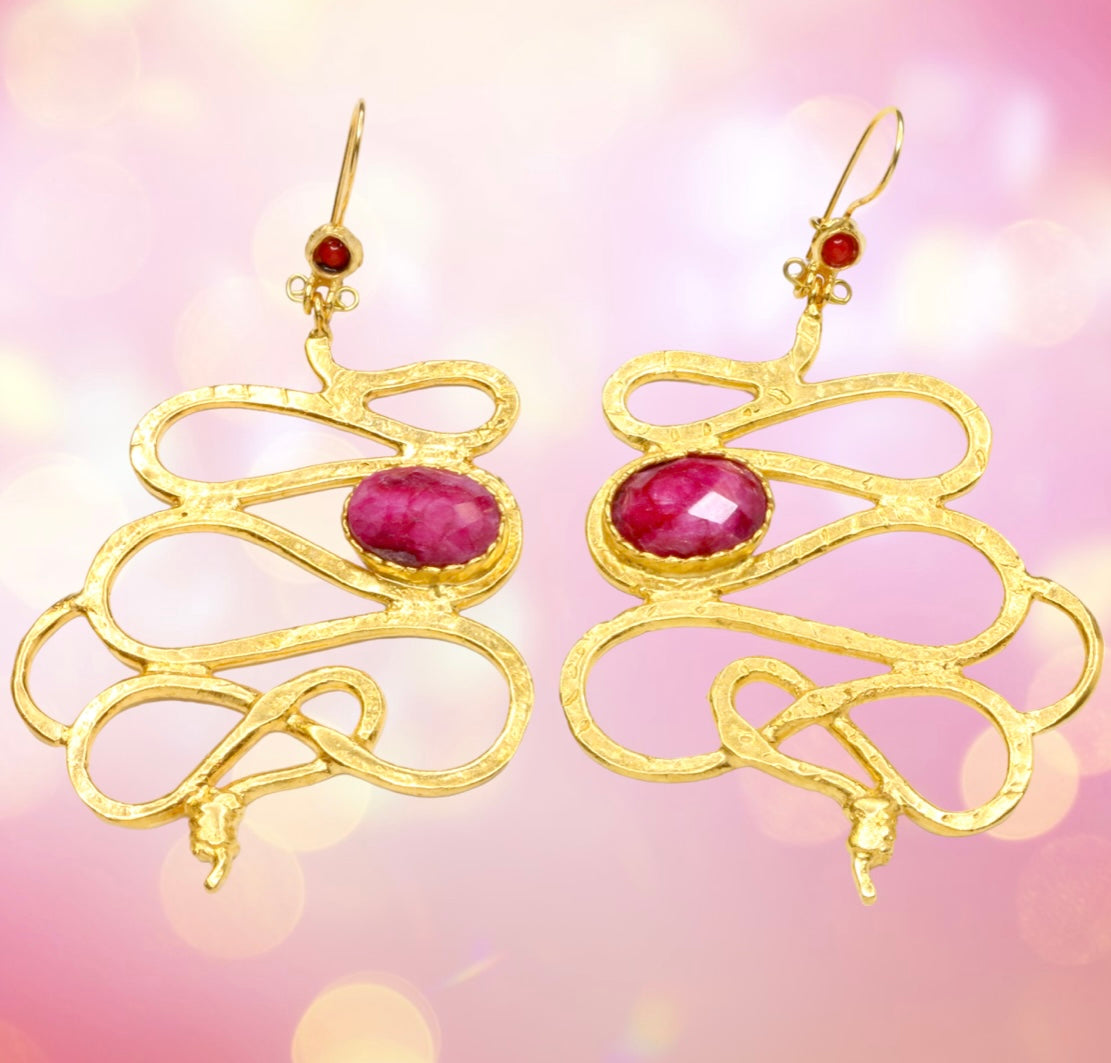 Gold colored snake shaped earrings with dark magenta stones on a pink background
