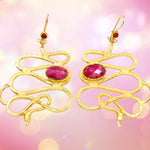 Gold colored snake shaped earrings with dark magenta stones on a pink background