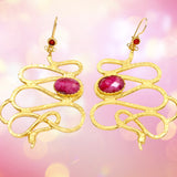 Gold colored snake shaped earrings with dark magenta stones on a pink background
