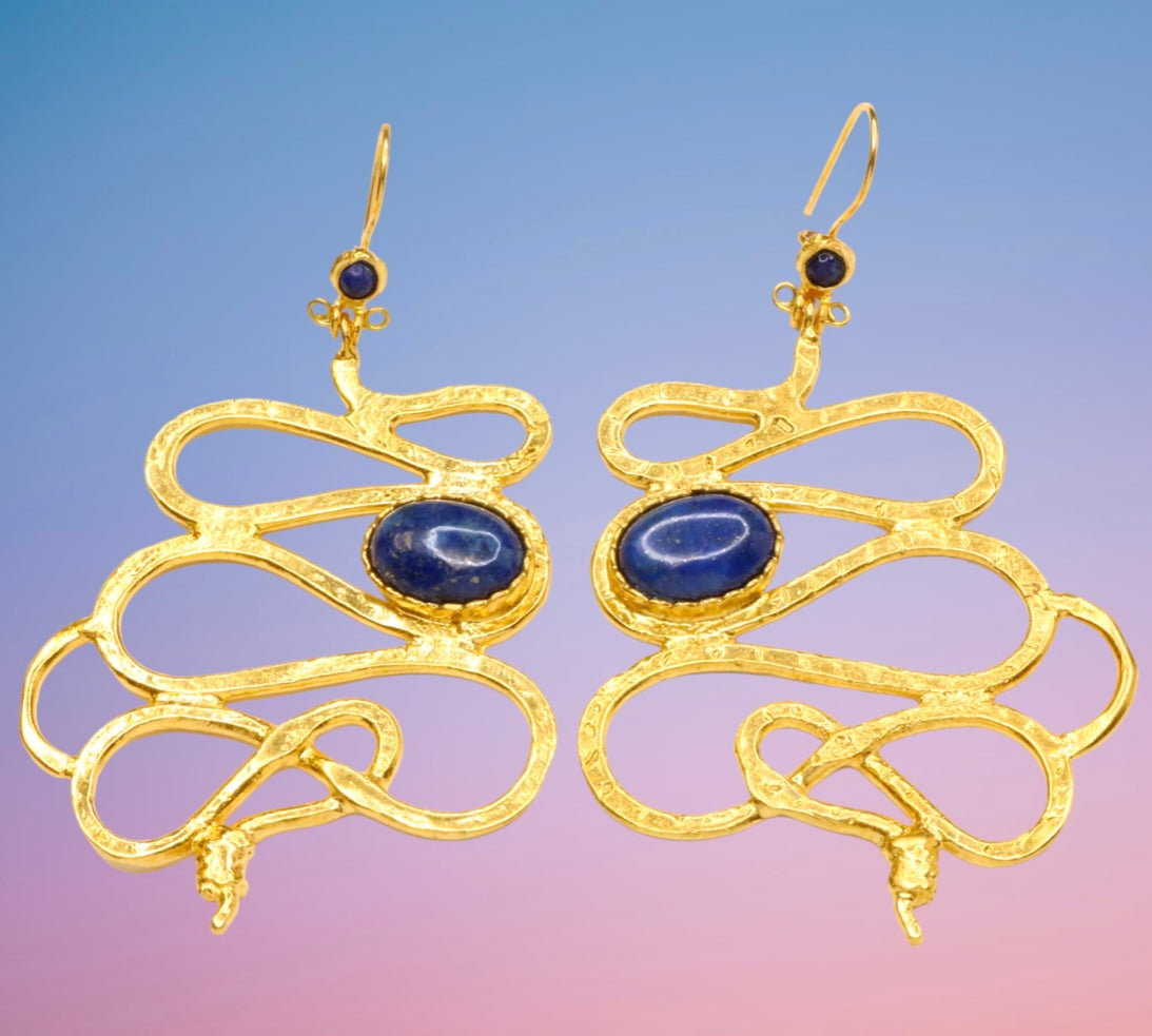 Gold colored snake shaped earrings with blue stones on a blue and pink background