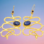 Gold colored snake shaped earrings with blue stones on a blue and pink background