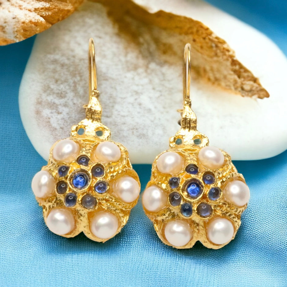 Gold flower shaped earrings with pearl like beads and blue stones on a blue background with white stone