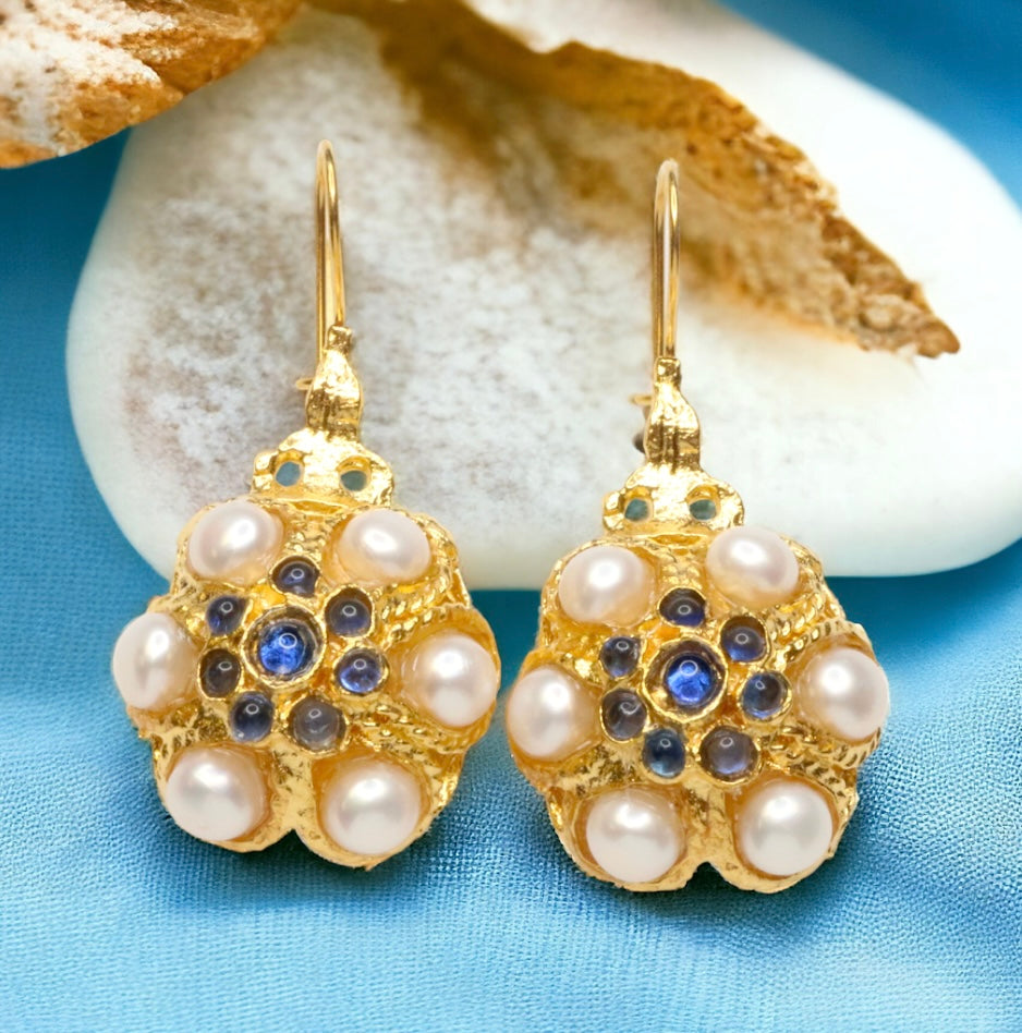 Gold flower shaped earrings with pearl like beads and blue stones on a blue background with white stone