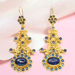Gold colored earrings with blue stone accents on a pink fabric background