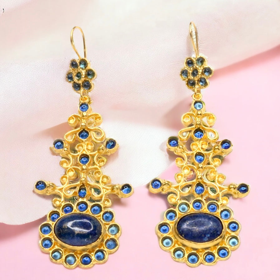 Gold colored earrings with blue stone accents on a pink fabric background