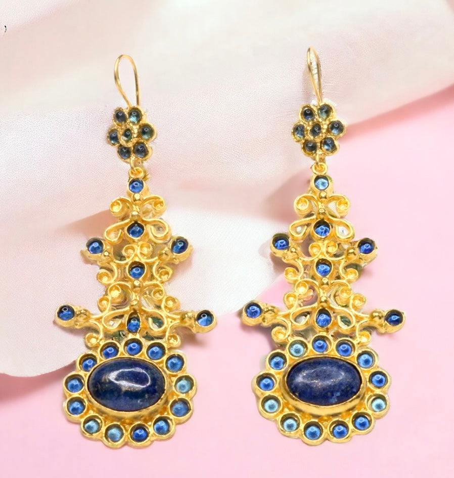 Gold colored earrings with blue stone accents on a pink fabric background