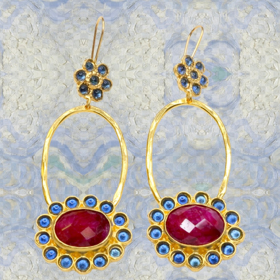 Gold colored earrings with dark magenta and blue stone accents on a blue painted background