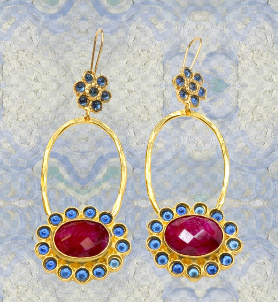 Gold colored earrings with dark magenta and blue stone accents on a blue painted background