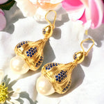 Gold colored earrings with pearl like accent and blue crystals on a white fabric background with flowers