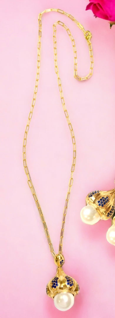 Gold colored necklace with a pearl like pendant and blue stone accents on a chain on a pink background