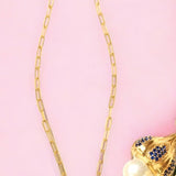Gold colored necklace with a pearl like pendant and blue stone accents on a chain on a pink background