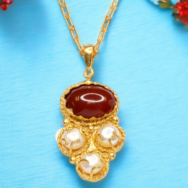 Gold colored necklace with a red stone and pearl-like accents on a gold elongated link chain on a blue background