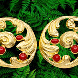 Pair of gold colored earrings with red stones accents on a green leaf background