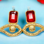 Gold colored eye shaped earrings with red crystal accents on a light blue background