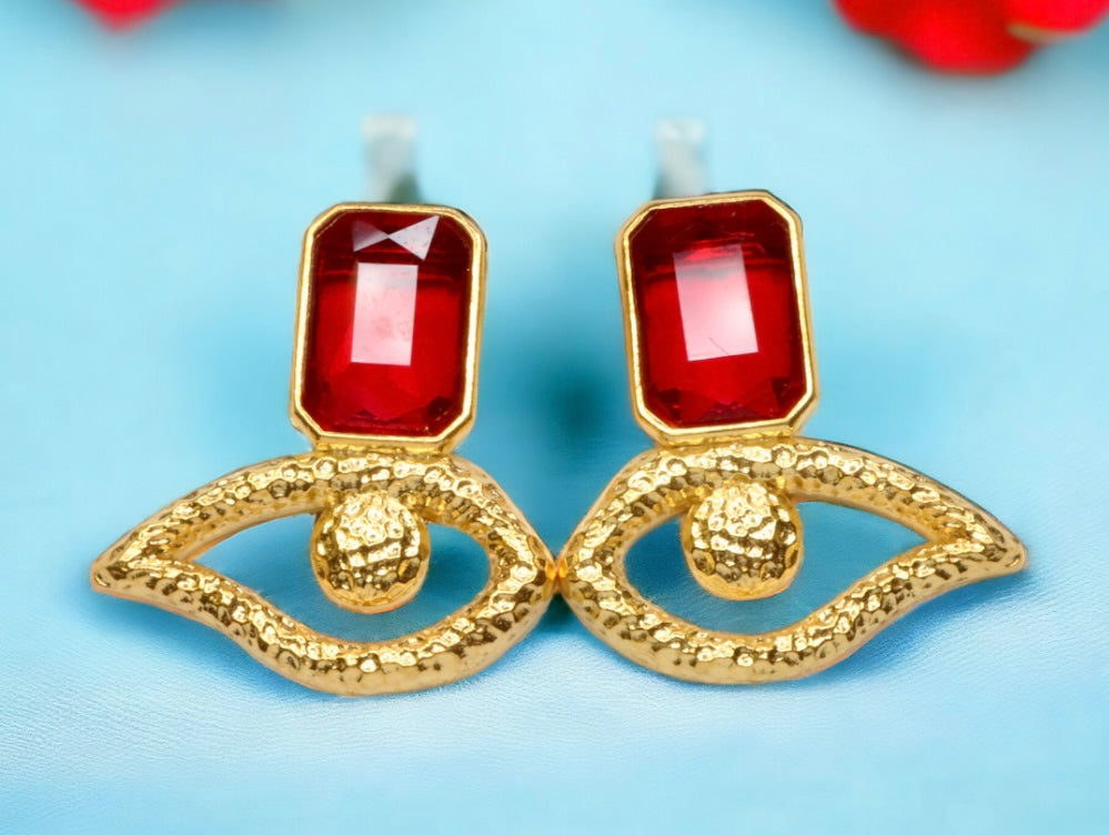 Gold colored eye shaped earrings with red crystal accents on a light blue background