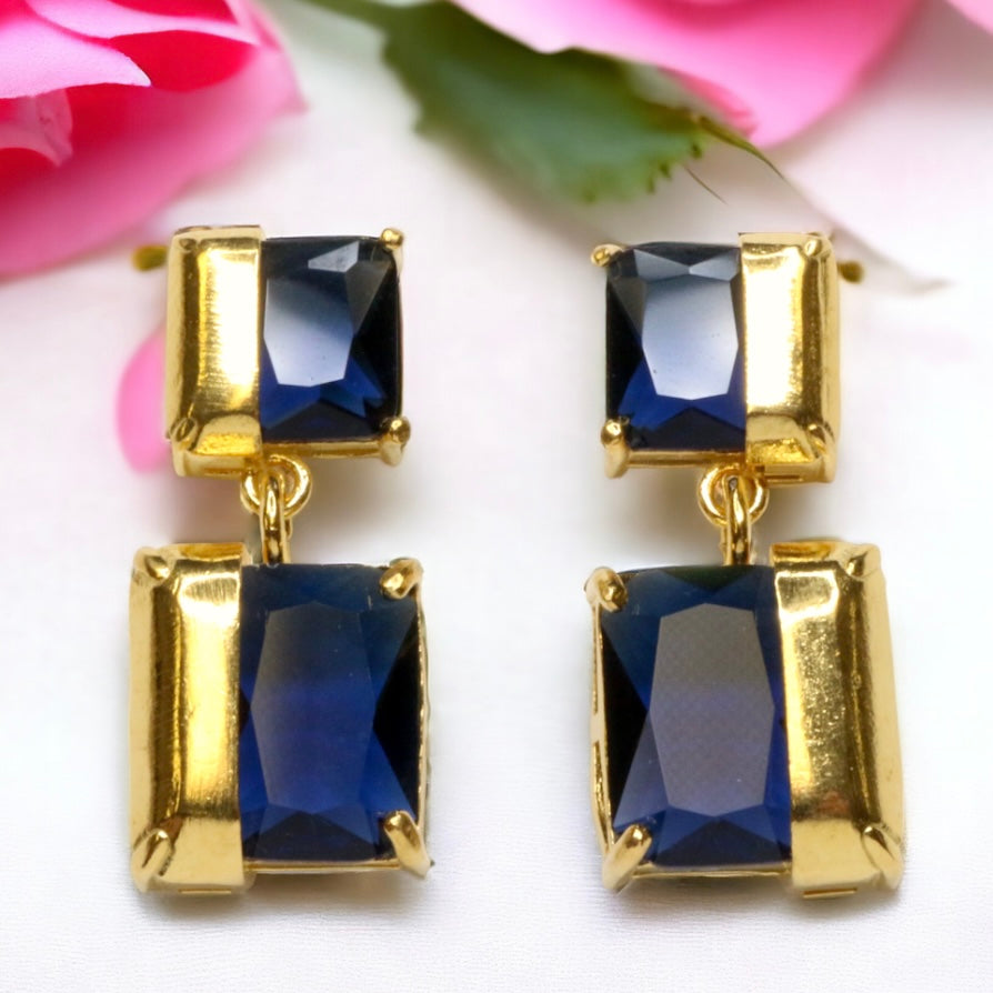 Gold colored earrings with square blue stones on a white background with pink flowers