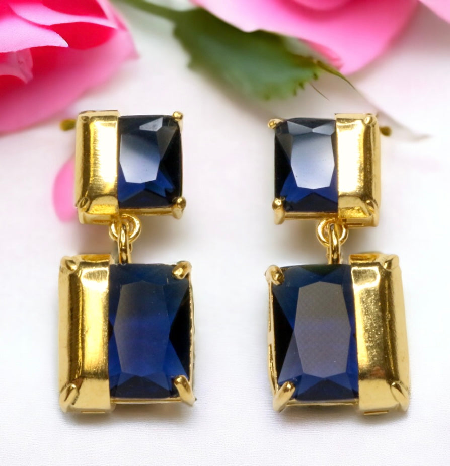 Gold colored earrings with square blue stones on a white background with pink flowers