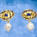 Gold colored eye shaped earrings with blue stones and pearl like accent drops on a blue background