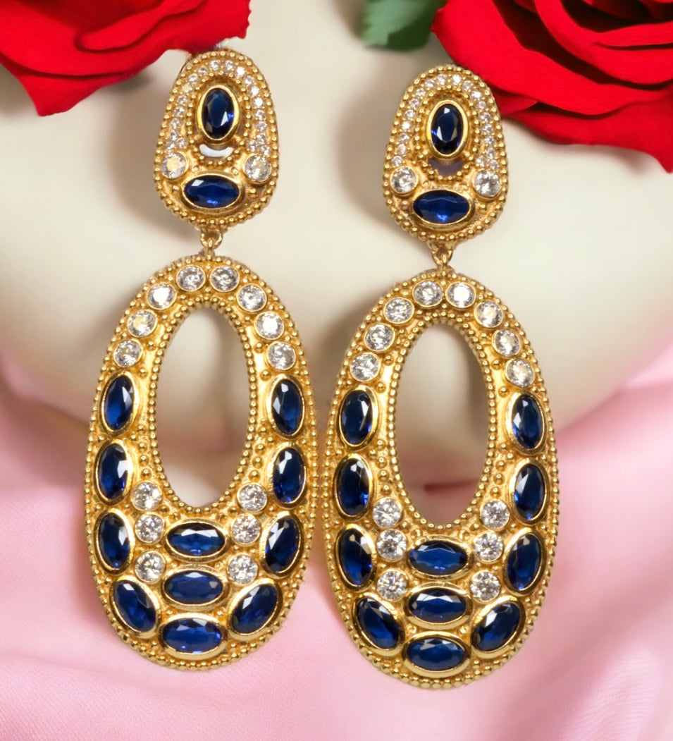 Gold colored earrings with dark blue and white crystal accents on a pink and white background