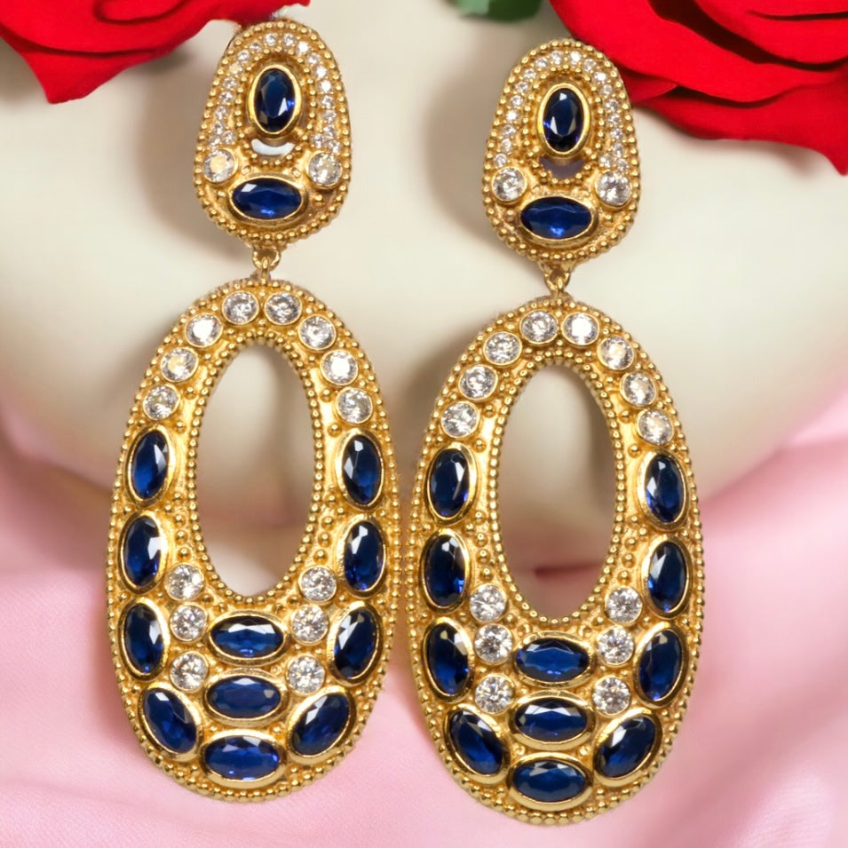 Gold colored earrings with dark blue and white crystal accents on a pink and white background