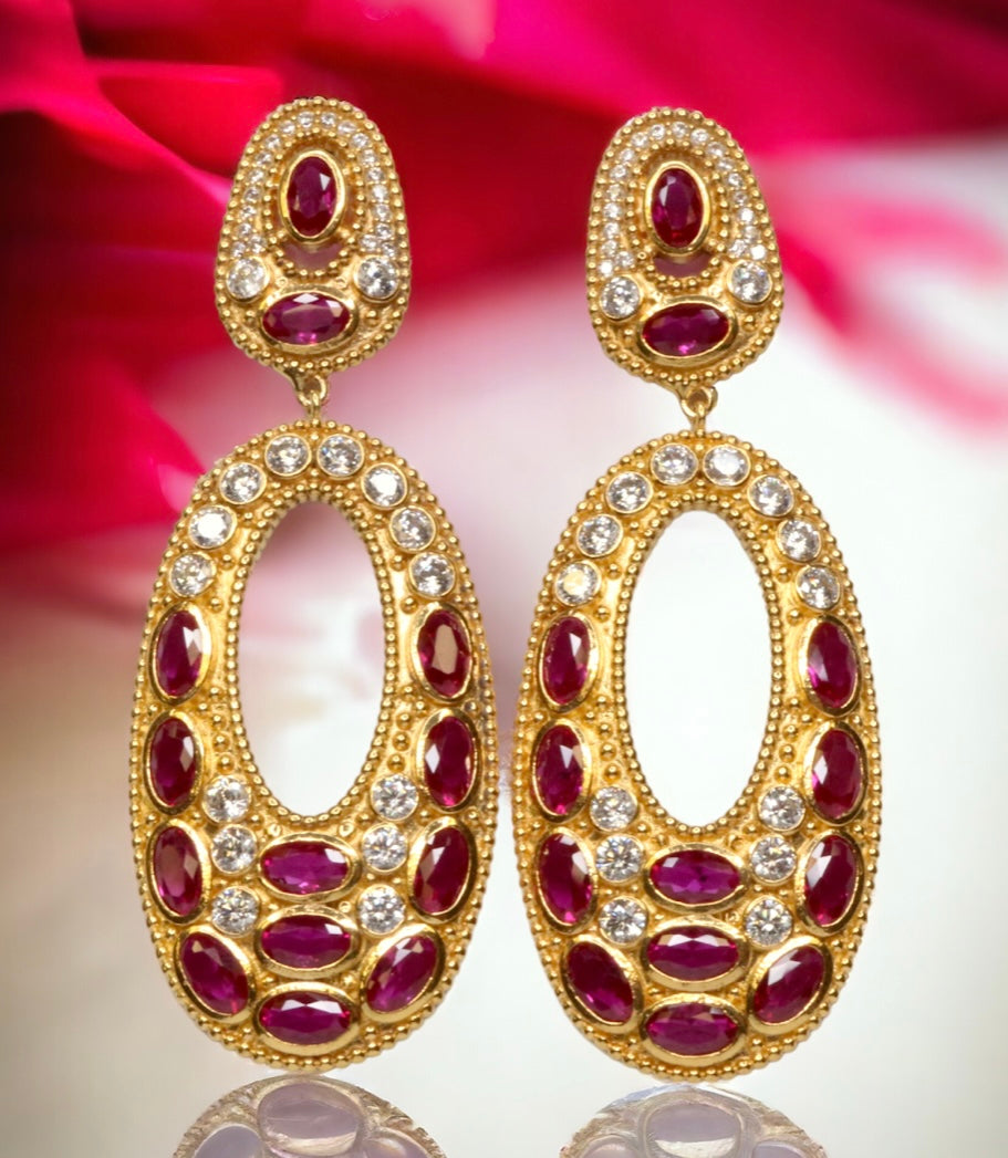 Gold colored earrings with dark magenta and white crystal accents on a red and white background