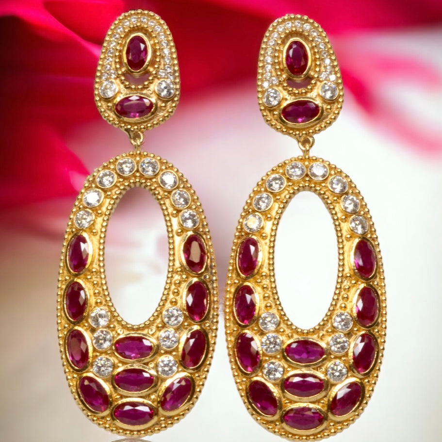 Gold colored earrings with dark magenta and white crystal accents on a red and white background