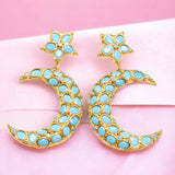 Gold and turquoise colored moon and star shaped earrings on a pink fabric background