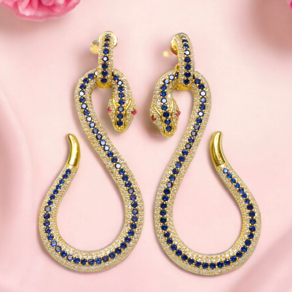 Gold snake-shaped earrings with blue and red crystals on a pink background