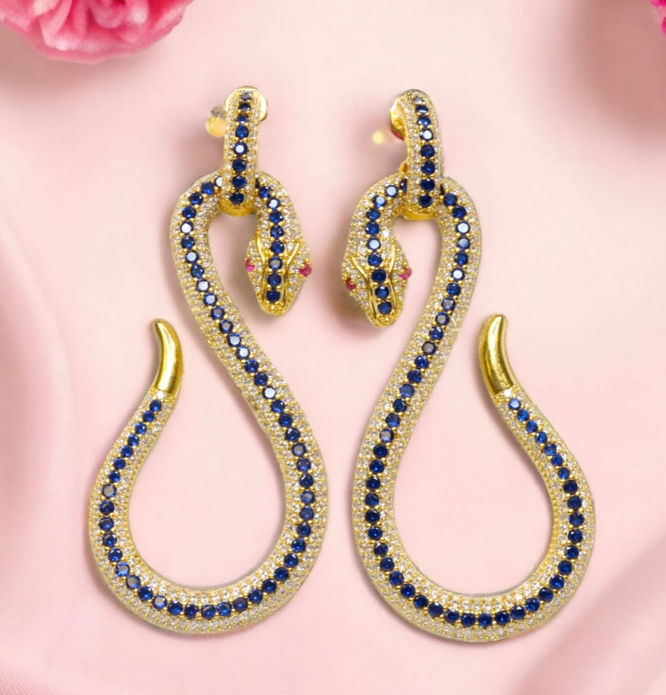 Gold snake-shaped earrings with blue and red crystals on a pink background