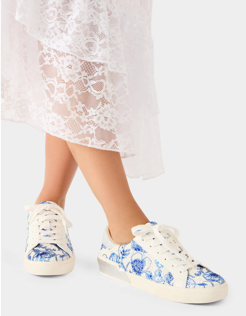 White sneakers with blue floral pattern worn by a person in a white lace dress.