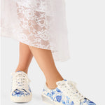 White sneakers with blue floral pattern worn by a person in a white lace dress.