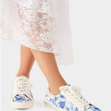 White sneakers with blue floral pattern worn by a person in a white lace dress.