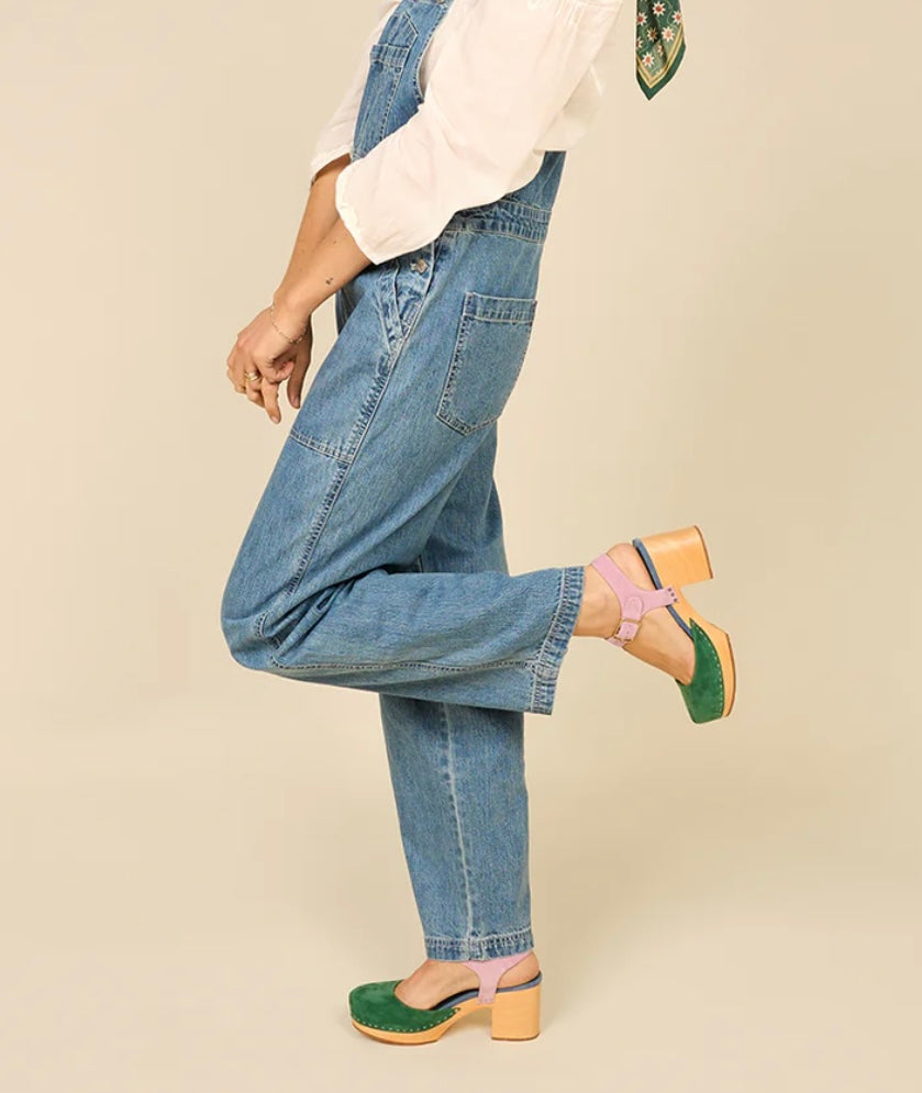 Green and blue suede clog shoe with pink strap with gold strap, and wooden sole pair on model standing with leg kicked up